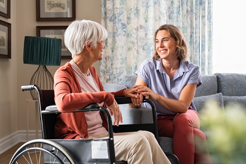 An older woman in a wheelchair smiling and holding hands with a younger woman at home